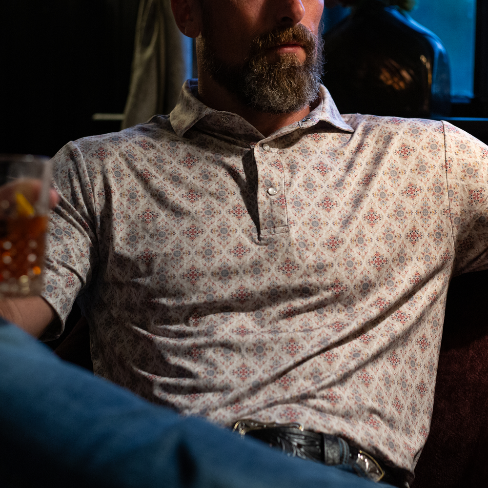 Man wearing The Classic Pearl Snap Polo by Iron Oak Apparel, showcasing an intricate floral western pattern with vibrant colors. He is seated indoors, holding a drink, embodying relaxed style and Texan authenticity.