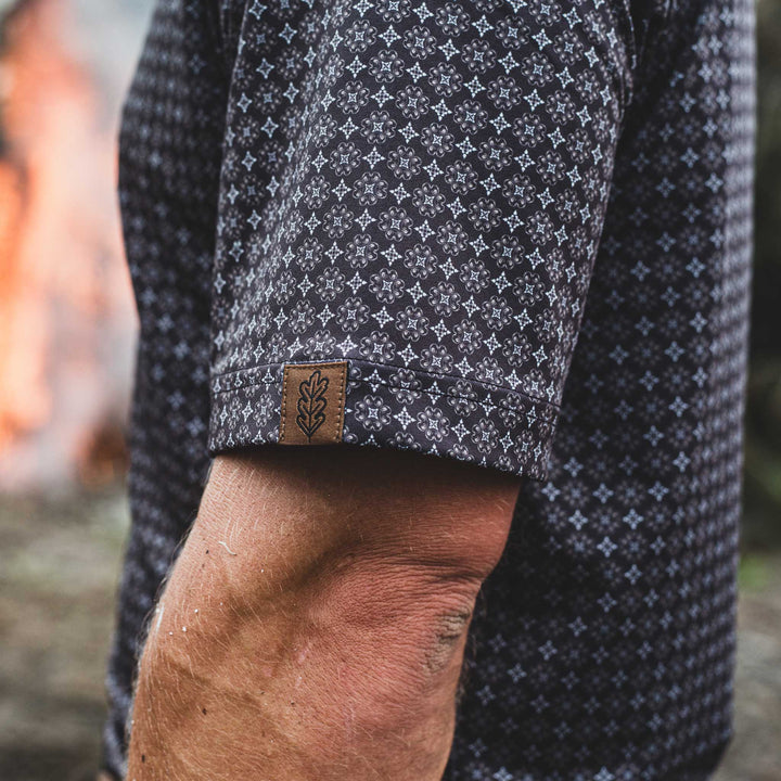 Close-up of a man wearing The Bandit Pearl Snap Polo by Iron Oak Apparel, featuring a detailed western pattern and the brand's logo on the sleeve. The stylish shirt combines modern design with Texan authenticity, perfect for any casual outfit.