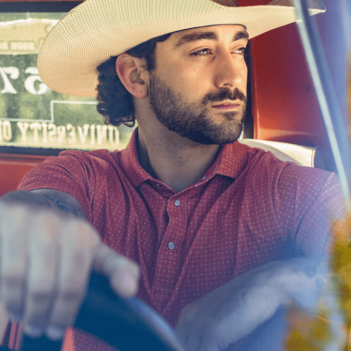 Man wearing The Boone Pearl Snap Polo by Iron Oak Apparel, featuring a burnt orange western pattern. He is sitting in a vintage truck, wearing a cowboy hat, capturing a blend of modern style and Texan authenticity. Perfect for a casual, rugged look.