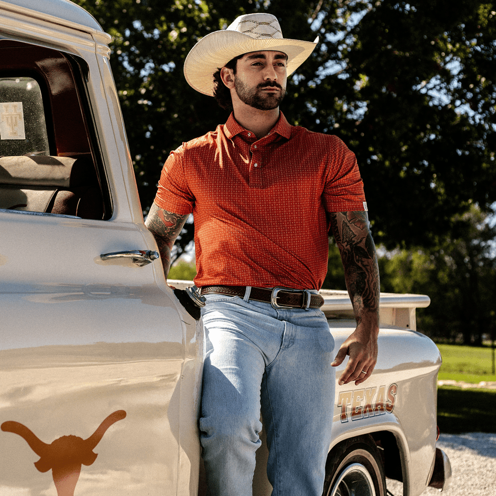 Man wearing The Boone Pearl Snap Polo by Iron Oak Apparel, featuring a burnt orange western pattern. He is leaning against a vintage Texas-themed truck, wearing a cowboy hat and light blue jeans, embodying modern Texan style and authenticity.
