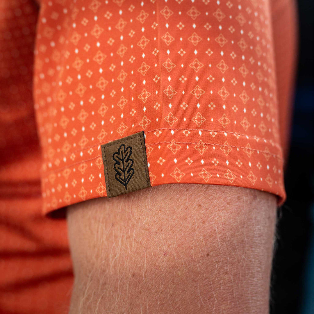 Close-up of the sleeve of The Boone Pearl Snap Polo by Iron Oak Apparel, featuring a burnt orange western pattern and the brand's logo. This stylish shirt blends modern design with Texan authenticity, perfect for any casual or dressy occasion.