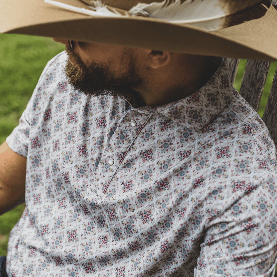 Man wearing The Classic Pearl Snap Polo by Iron Oak Apparel, showcasing an intricate floral western pattern with vibrant colors. He is seated outdoors, wearing a cowboy hat with feathers, embodying relaxed style and Texan authenticity. Perfect for casual and social settings.