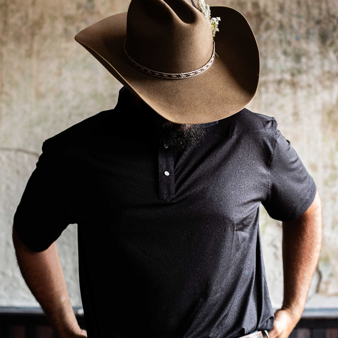 Man wearing The Folsom Western Yoke Pearl Snap Polo by Iron Oak Apparel, featuring a subtle black paisley Western pattern. He stands with a cowboy hat, exuding a rugged yet refined style.