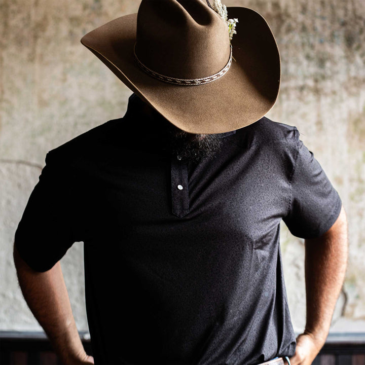 Man wearing The Folsom Western Yoke Pearl Snap Polo by Iron Oak Apparel, featuring a subtle black paisley Western pattern. He stands with a cowboy hat, exuding a rugged yet refined style.
