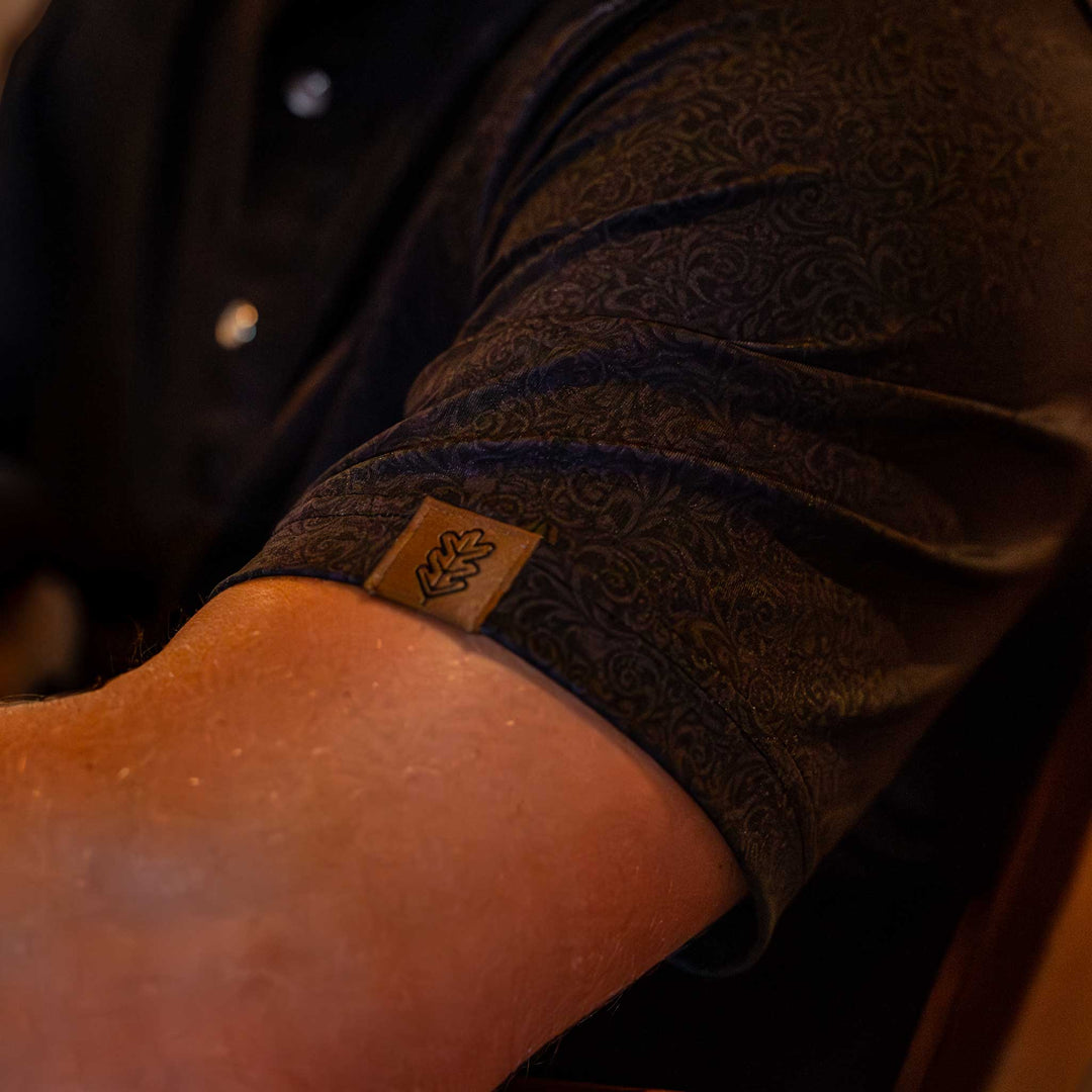 Close-up of the sleeve of The Folsom Western Yoke Pearl Snap Polo by Iron Oak Apparel, featuring a subtle black paisley Western pattern. The sleeve has the Iron Oak logo tag, adding a touch of authenticity.
