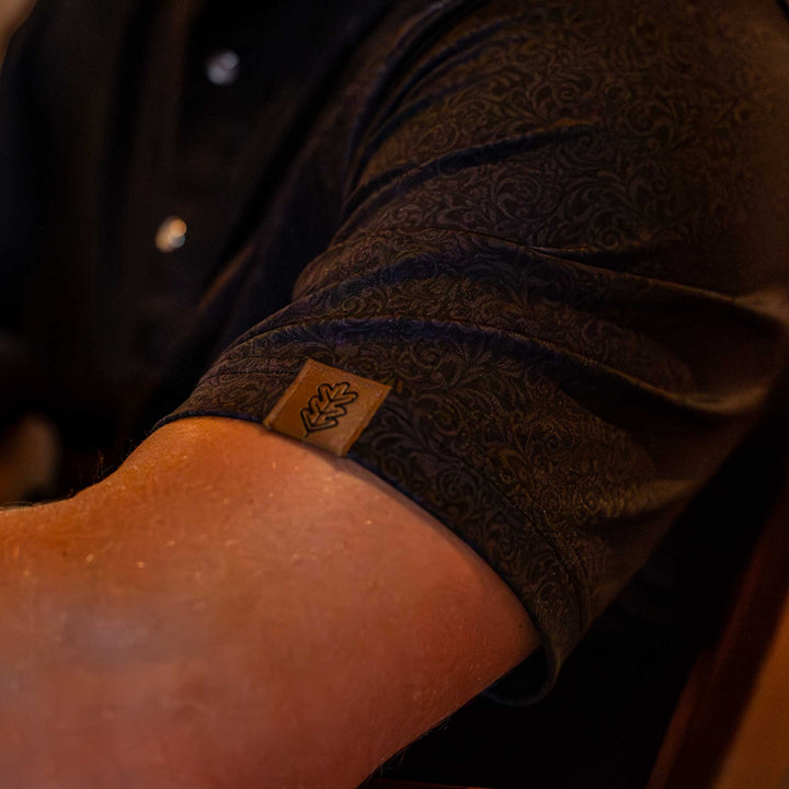 Close-up of the sleeve of The Folsom Western Yoke Pearl Snap Polo by Iron Oak Apparel, featuring a subtle black paisley Western pattern. The sleeve has the Iron Oak logo tag, adding a touch of authenticity.