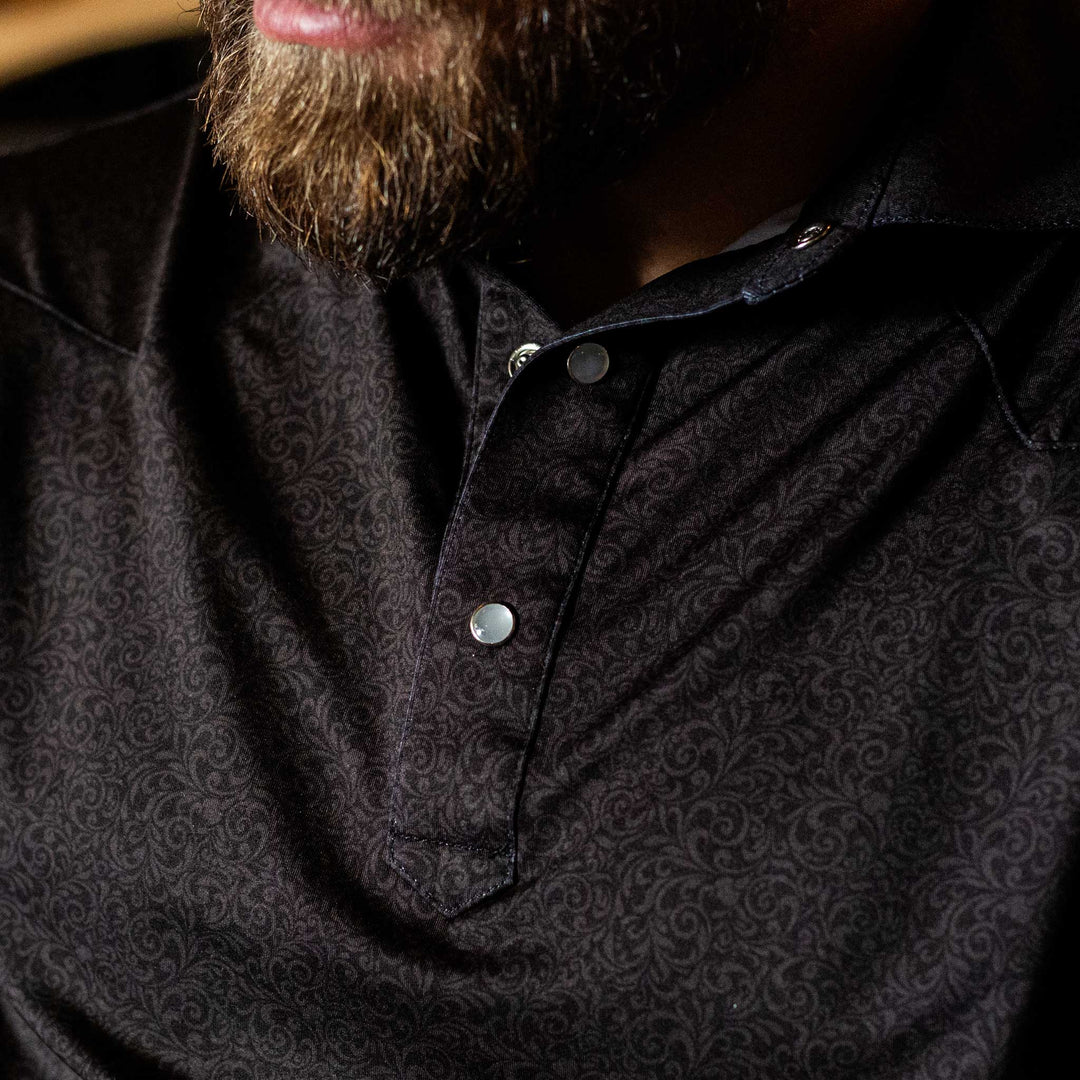 Man wearing The Folsom Western Yoke Pearl Snap Polo by Iron Oak Apparel, featuring a subtle black paisley Western pattern. The close-up shows the intricate design and pearl snap buttons.