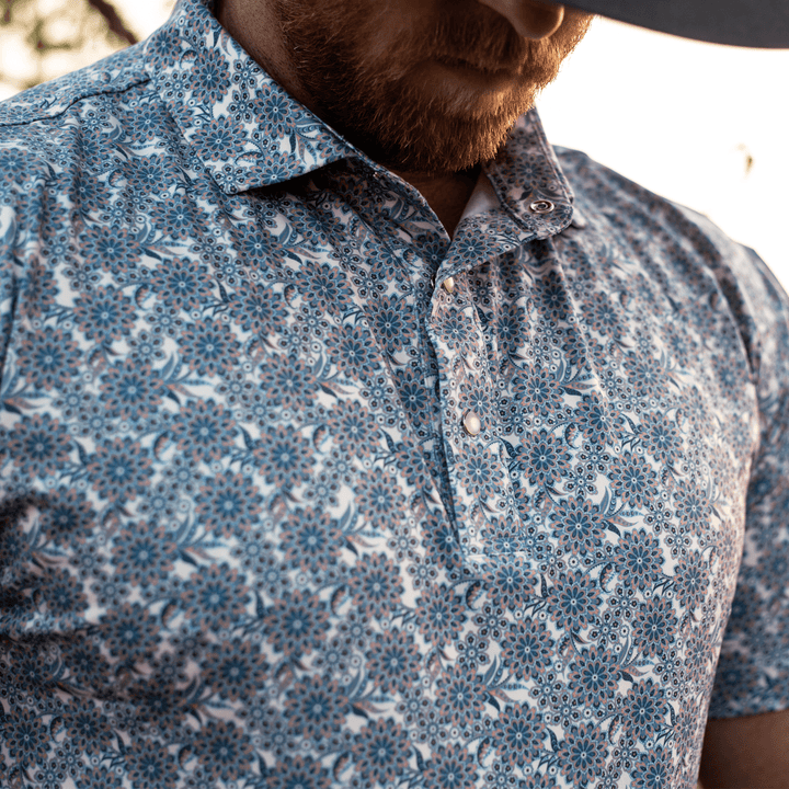 Man wearing The Gentry Western Yoke Pearl Snap Polo by Iron Oak Apparel, showcasing the vibrant blue floral pattern. He stands outdoors, embodying a classic yet modern Western look.