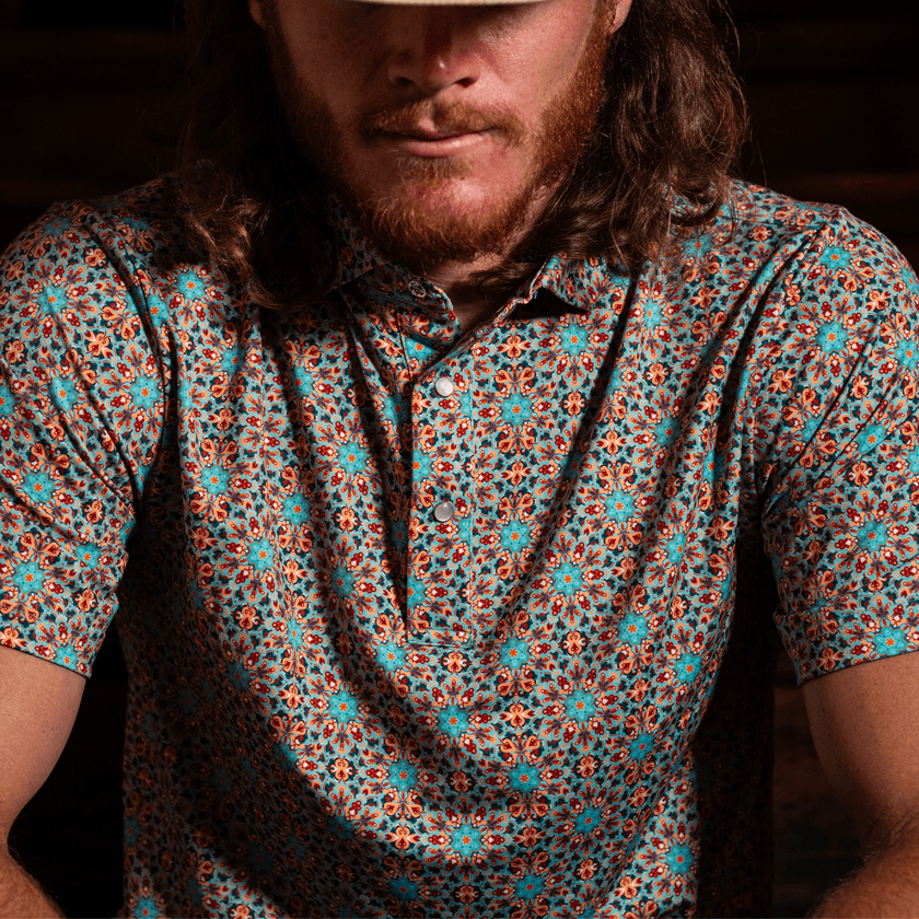 Man wearing The Clementine Pearl Snap Polo by Iron Oak Apparel, featuring a vibrant geometric western pattern with turquoise and orange accents. He is sitting indoors with a focused expression, wearing a cowboy hat, embodying relaxed style and Texan authenticity. Perfect for casual and social settings.