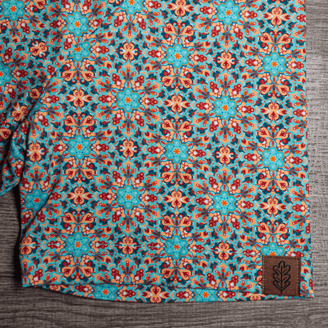 Close-up of the hem of The Clementine Pearl Snap Polo by Iron Oak Apparel, showcasing the vibrant geometric western pattern with turquoise and orange accents and the brand's logo. This stylish shirt blends modern design with Texan authenticity, ideal for any occasion.