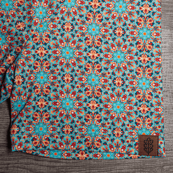 Close-up of the hem of The Clementine Pearl Snap Polo by Iron Oak Apparel, showcasing the vibrant geometric western pattern with turquoise and orange accents and the brand's logo. This stylish shirt blends modern design with Texan authenticity, ideal for any occasion.