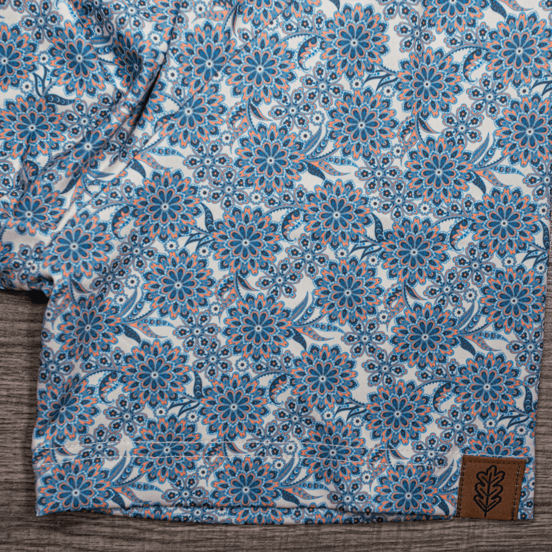 Detailed close-up of the sleeve of The Gentry Western Yoke Pearl Snap Polo by Iron Oak Apparel, showcasing the vibrant blue floral pattern and the Iron Oak logo tag.