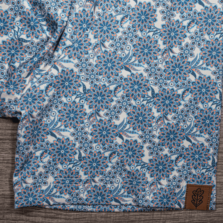 Detailed close-up of the sleeve of The Gentry Western Yoke Pearl Snap Polo by Iron Oak Apparel, showcasing the vibrant blue floral pattern and the Iron Oak logo tag.