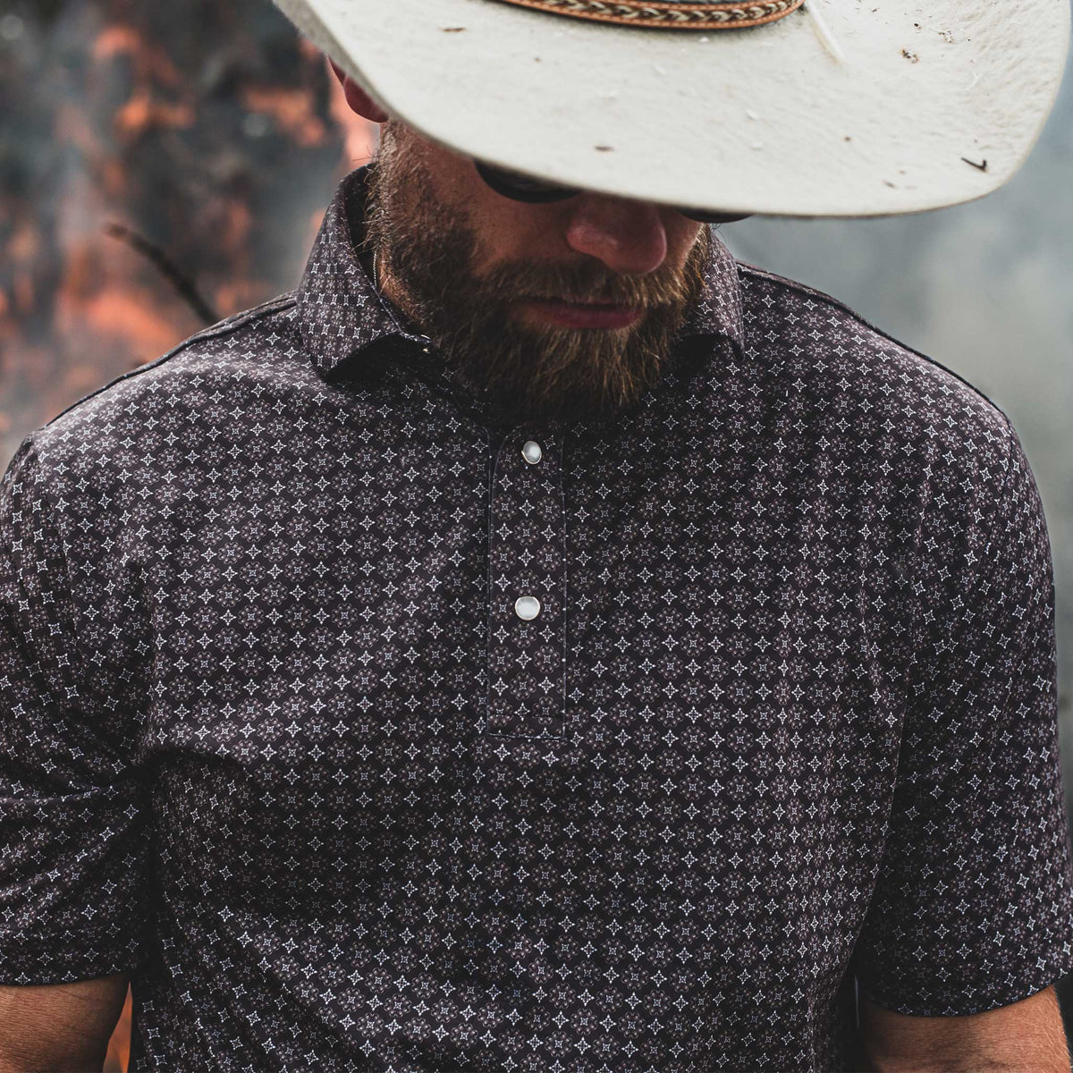Man wearing The Bandit Pearl Snap Polo by Iron Oak Apparel, with a white cowboy hat and sunglasses. The shirt features a detailed western pattern, blending modern style with Texan authenticity. Perfect for a rugged yet stylish look.