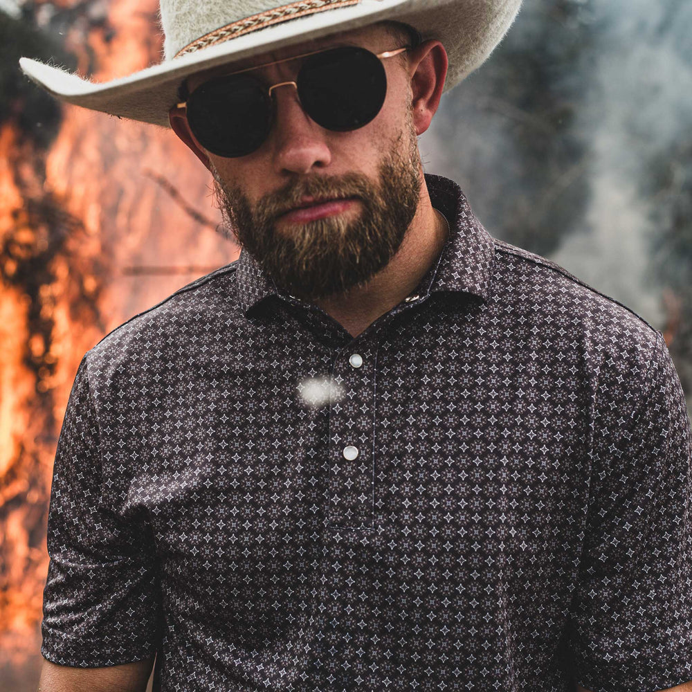 Man wearing The Bandit Pearl Snap Polo by Iron Oak Apparel, featuring a refined western pattern. He pairs it with a cowboy hat and sunglasses, standing against a dramatic background of flames and smoke. This stylish shirt embodies modern Texan fashion and rugged authenticity.