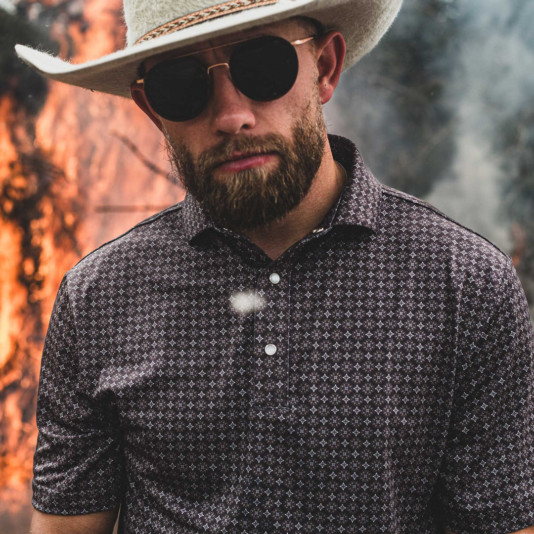  Man wearing The Bandit Pearl Snap Polo by Iron Oak Apparel, featuring a refined western pattern. He pairs it with a cowboy hat and sunglasses, standing against a dramatic background of flames and smoke. This stylish shirt embodies modern Texan fashion and rugged authenticity.