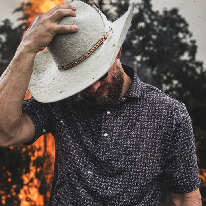 Man tipping his white cowboy hat, wearing The Bandit Pearl Snap Polo by Iron Oak Apparel. The shirt features a detailed western pattern, set against a dramatic backdrop of flames and smoke. This look embodies modern Texan style and rugged authenticity.