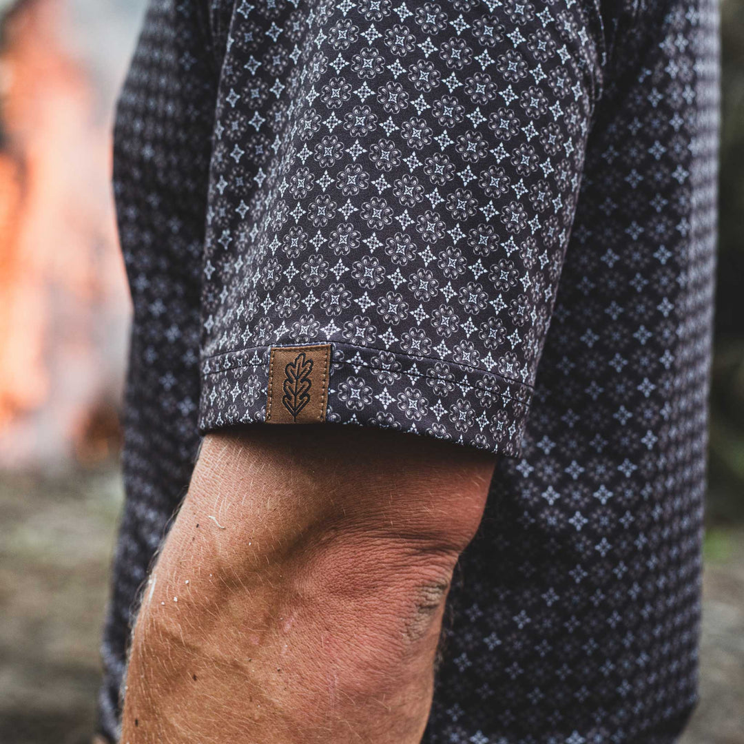  Close-up of a man wearing The Bandit Pearl Snap Polo by Iron Oak Apparel, featuring a detailed western pattern and the brand's logo on the sleeve. The stylish shirt combines modern design with Texan authenticity, perfect for any casual outfit.