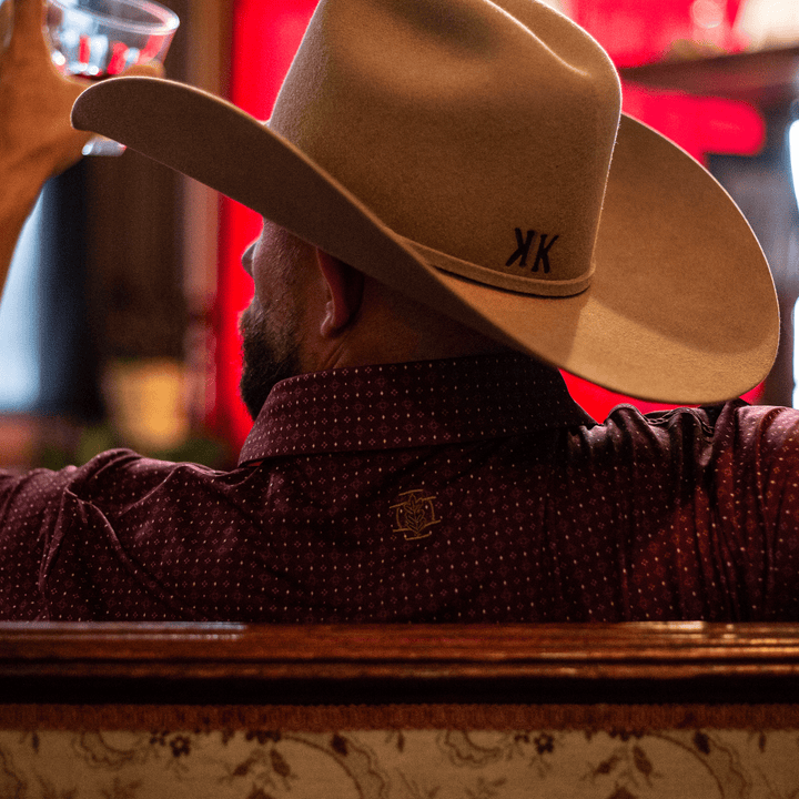 Man wearing The Bennett Pearl Snap Polo by Iron Oak Apparel, featuring a maroon western pattern and embroidered logo on the back. He is sitting in a bar, holding up a glass of whiskey, and wearing a beige cowboy hat. The scene captures modern Texan style and authenticity, perfect for a relaxed, stylish evening.