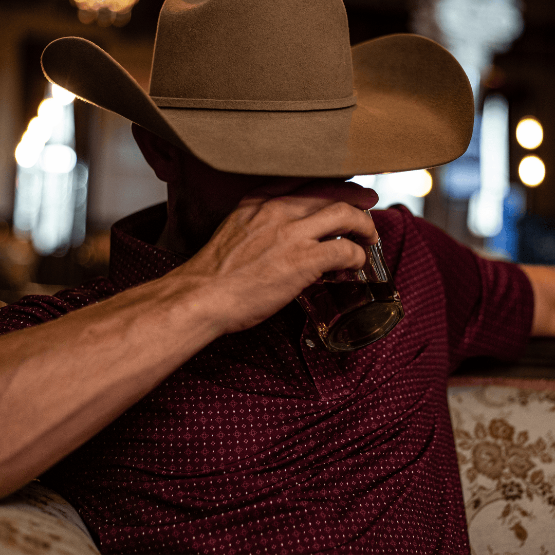 Man in a beige cowboy hat sipping whiskey, wearing The Bennett Pearl Snap Polo by Iron Oak Apparel. The maroon shirt features a western pattern, blending modern style with Texan authenticity, perfect for a relaxed evening out.