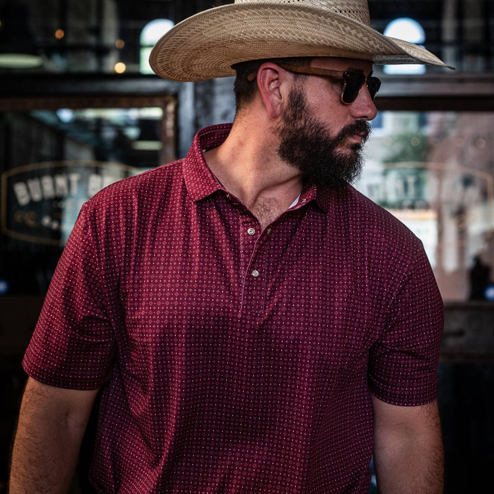 Man wearing The Bennett Pearl Snap Polo by Iron Oak Apparel, featuring a rich maroon western pattern. He pairs it with a straw cowboy hat and sunglasses, capturing a blend of modern style and Texan authenticity. Perfect for casual outings or a night out.