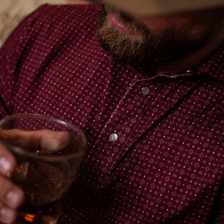 Close-up of a man wearing The Bennett Pearl Snap Polo by Iron Oak Apparel, featuring a maroon western pattern. He is holding a glass of whiskey, capturing a blend of modern style and Texan authenticity. Perfect for relaxed and stylish occasions.