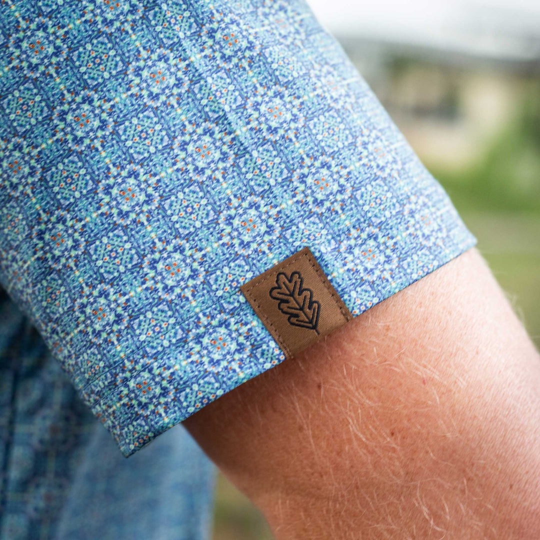 Close-up of the sleeve of The Boot Scoot Pearl Snap Polo by Iron Oak Apparel, featuring a vibrant blue western pattern and the brand's logo. 