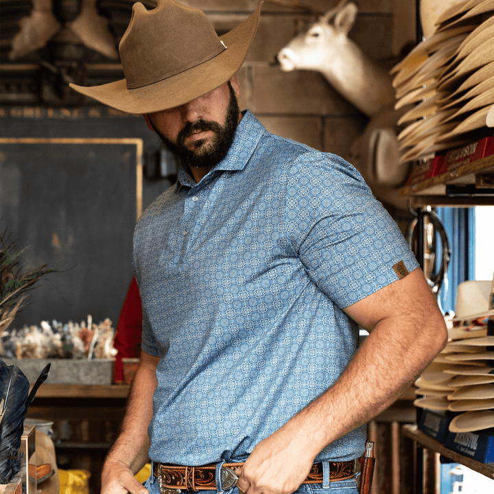 Man wearing The Boot Scoot Pearl Snap Polo by Iron Oak Apparel, featuring a vibrant blue western pattern. He pairs it with a brown cowboy hat and belt, embodying modern Texan style and authenticity