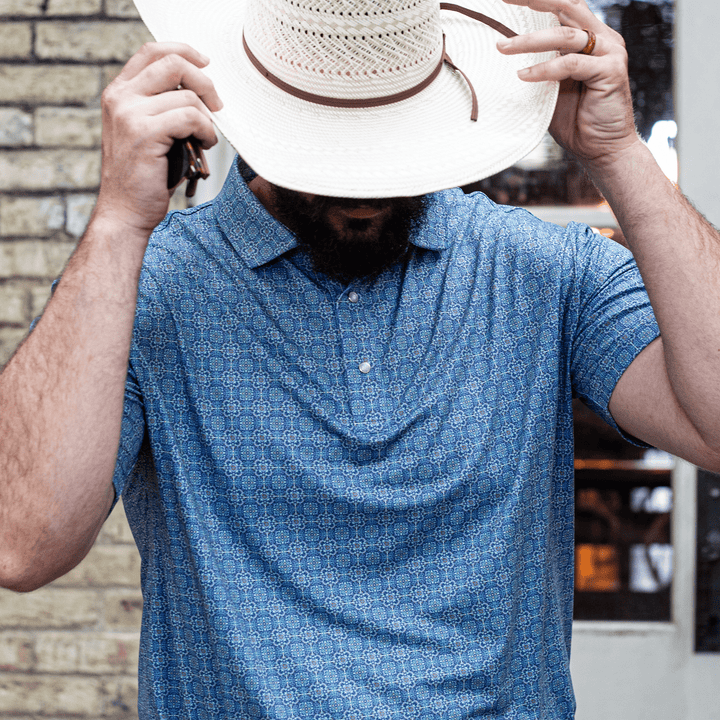 Man wearing The Boot Scoot Pearl Snap Polo by Iron Oak Apparel, featuring a vibrant blue western pattern. He is adjusting his straw cowboy hat, capturing a blend of modern style and Texan authenticity. Perfect for casual outings or a night out.