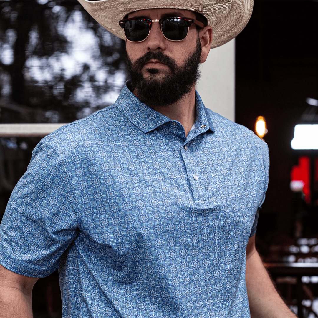 Man wearing The Boot Scoot Pearl Snap Polo by Iron Oak Apparel, featuring a vibrant blue western pattern. He pairs it with a straw cowboy hat and sunglasses, embodying modern Texan style and authenticity.
