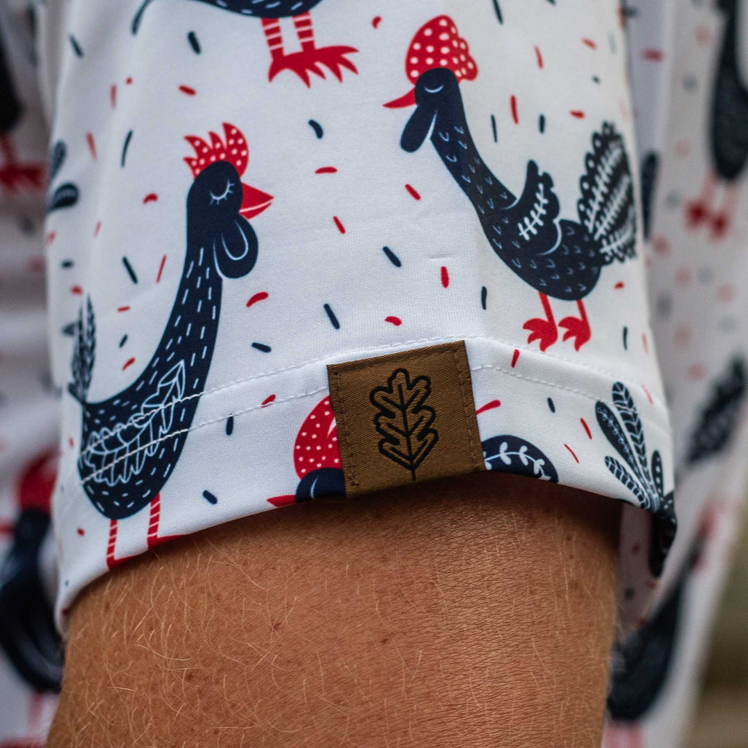 Close-up of the sleeve of The Cock-a-doo Pearl Snap Polo by Iron Oak Apparel, showcasing the whimsical pattern of navy blue roosters with red accents on a white background and the brand's logo. This fun and stylish shirt is ideal for adding personality to any outfit.