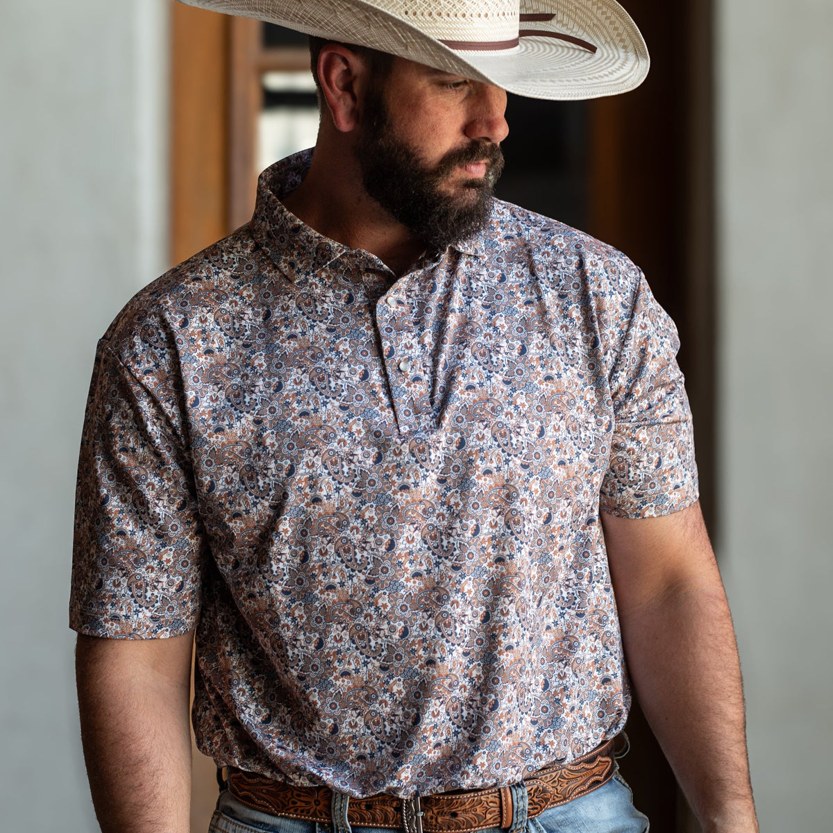 Man wearing The Dosey Doe Pearl Snap Polo by Iron Oak Apparel, featuring a vibrant paisley Western pattern in shades of orange, blue, and white. He is standing with a straw hat, embodying a rugged yet stylish look.