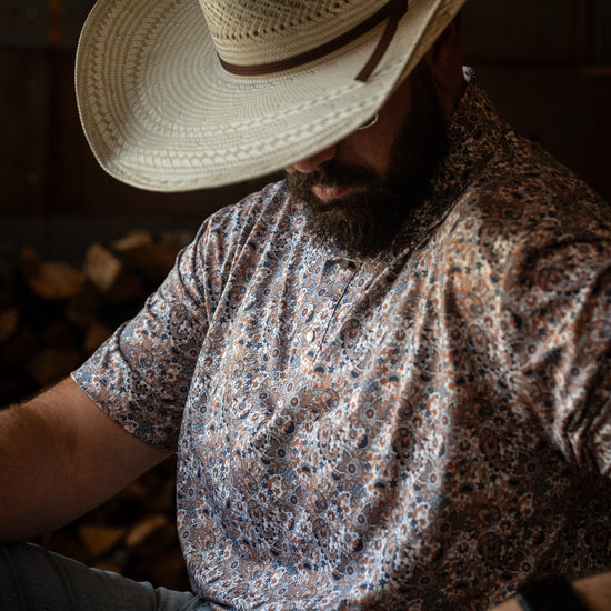 Man wearing The Dosey Doe Pearl Snap Polo by Iron Oak Apparel, featuring a vibrant paisley Western pattern in shades of orange, blue, and white. He is seated with a straw hat, showcasing a blend of rugged and stylish.