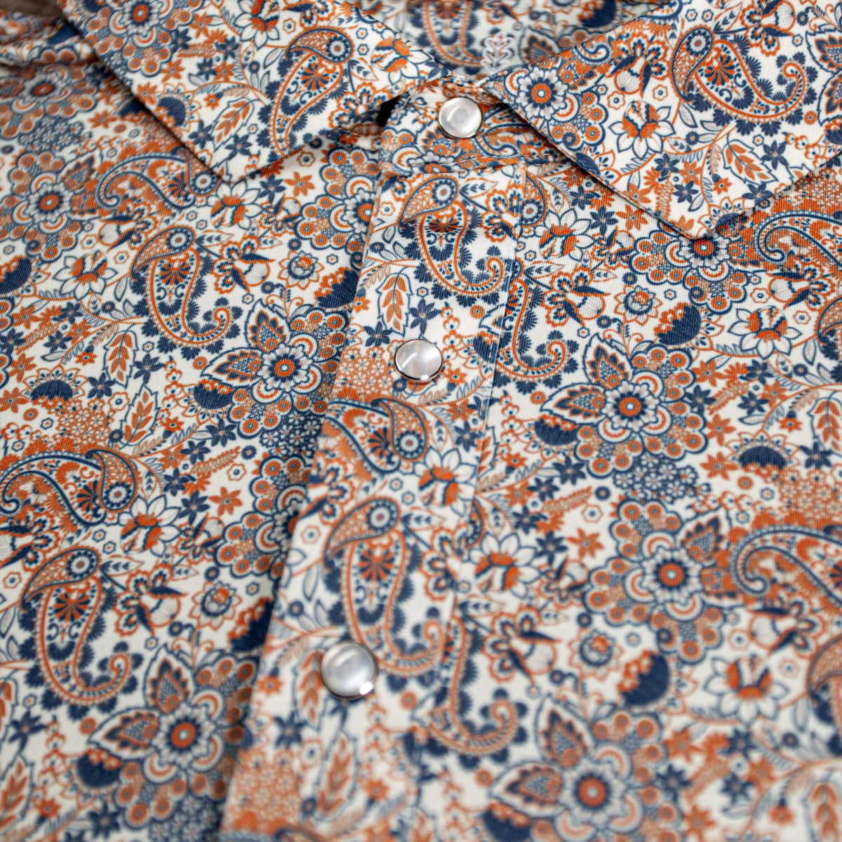Close-up of The Dosey Doe Pearl Snap Polo by Iron Oak Apparel, highlighting the vibrant paisley Western pattern in shades of orange, blue, and white. The shirt features signature pearl snap buttons, adding a bold statement.
