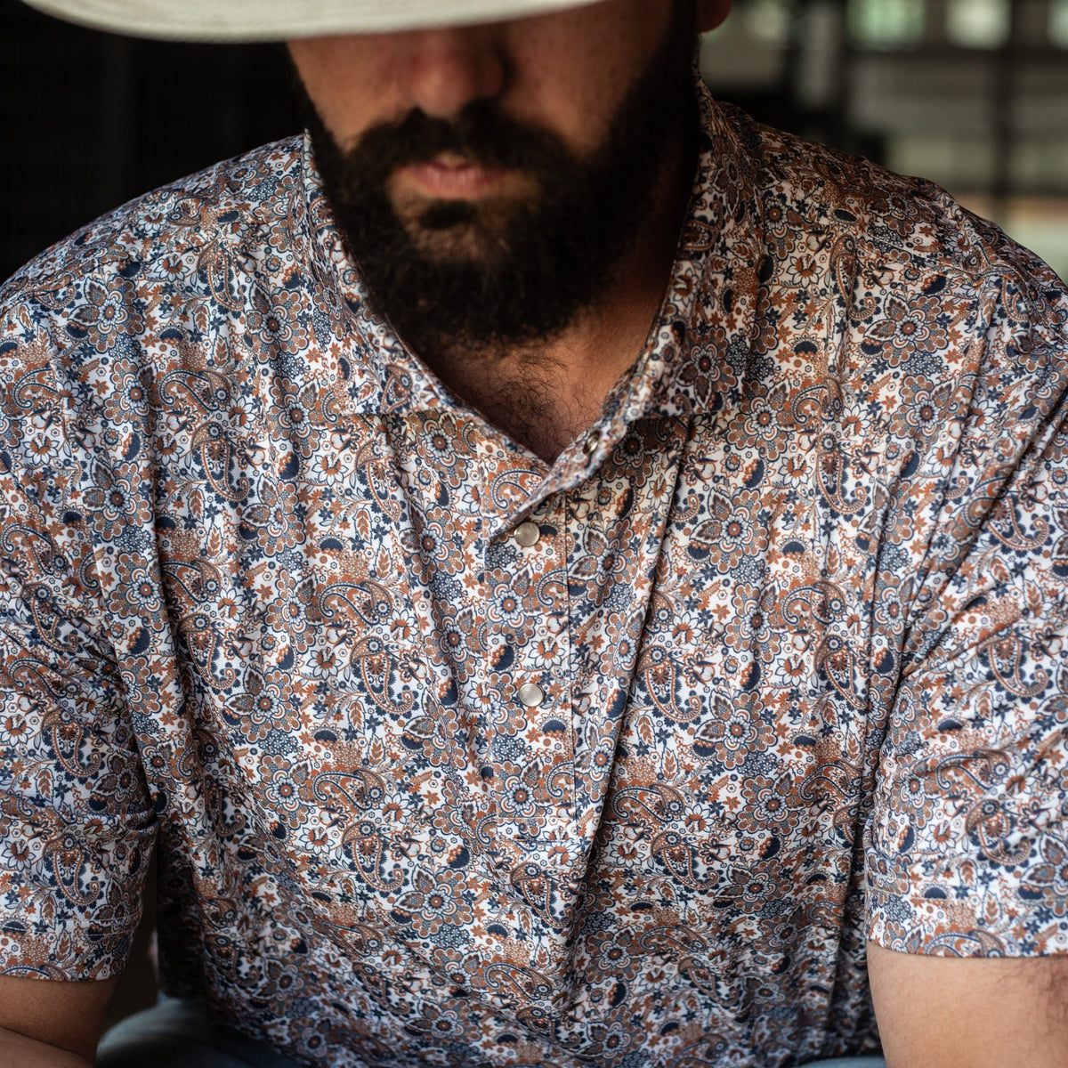 Man wearing The Dosey Doe Pearl Snap Polo by Iron Oak Apparel, featuring a vibrant paisley Western pattern in shades of orange, blue, and white. He is seated, looking down with a straw hat, showcasing a blend of rugged and stylish.