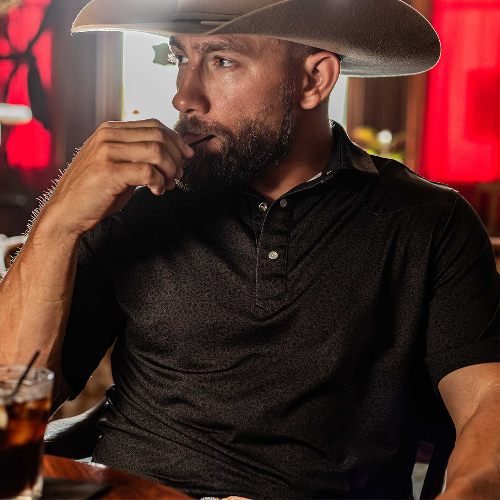  Man wearing The Folsom Western Yoke Pearl Snap Polo by Iron Oak Apparel, featuring a subtle black paisley Western pattern. He is seated, wearing a cowboy hat, adding a rugged yet refined touch.