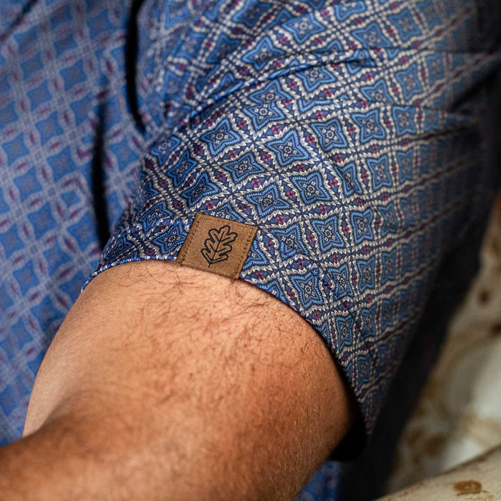 Detailed close-up of the sleeve of The Granger Western Yoke Pearl Snap Polo by Iron Oak Apparel, showcasing the blue Western pattern and the Iron Oak logo tag.