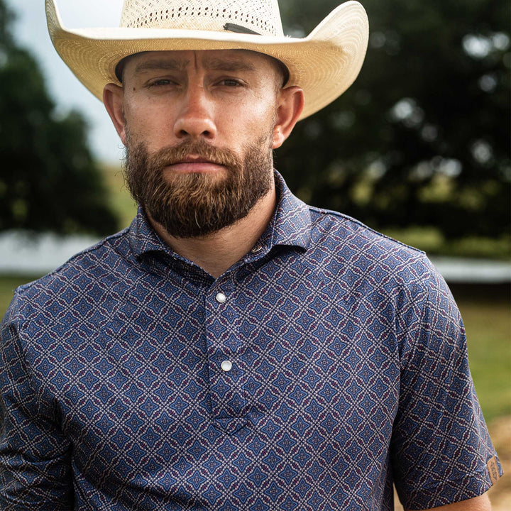 Man wearing The Granger Western Yoke Pearl Snap Polo by Iron Oak Apparel, highlighting the classic blue Western pattern. He stands outdoors, exuding confidence and style.