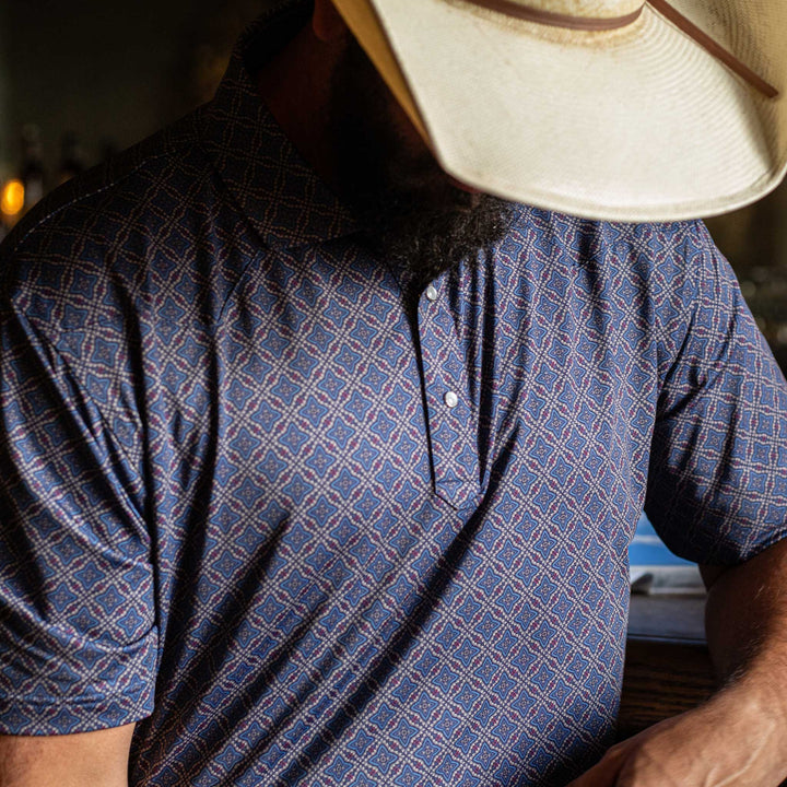 Man wearing The Granger Western Yoke Pearl Snap Polo by Iron Oak Apparel, showcasing the intricate blue Western pattern. He sits indoors, embodying a classic yet modern Western look.