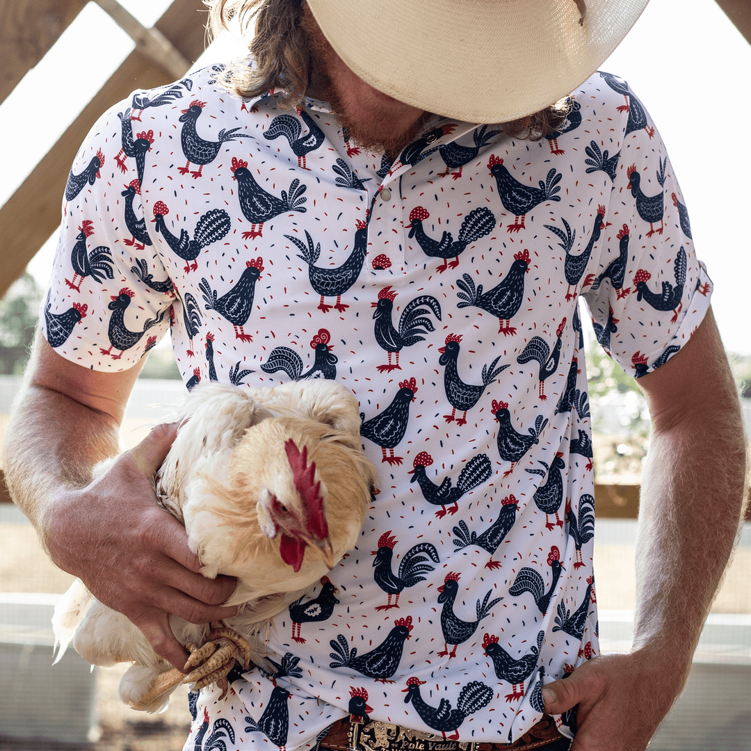 Man wearing The Cock-a-doo Pearl Snap Polo by Iron Oak Apparel, featuring a whimsical pattern of navy blue roosters with red accents on a white background. He is holding a live chicken and wearing a cowboy hat, embodying playful style and Texan authenticity.