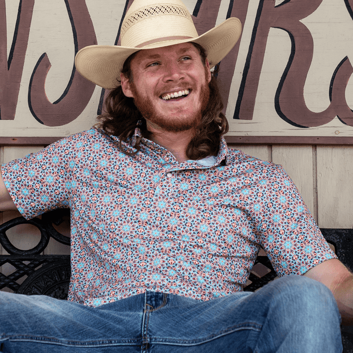 Man wearing The Clementine Pearl Snap Polo by Iron Oak Apparel, showcasing a vibrant geometric western pattern with turquoise and orange accents. He is sitting outside, smiling, and wearing a cowboy hat, embodying relaxed style and Texan authenticity. Perfect for casual and social settings.