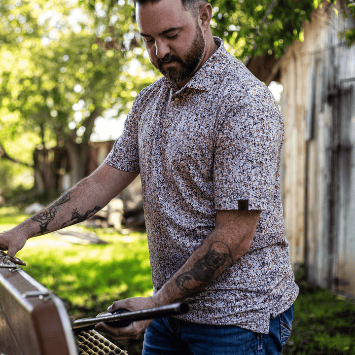 Man wearing the CamOAK Pearl Snap Polo by Iron Oak Apparel, showcasing a camouflage pattern with earthy tones. He is standing outdoors, holding a tool, embodying rugged style and Texan authenticity. Perfect for active, casual wear.