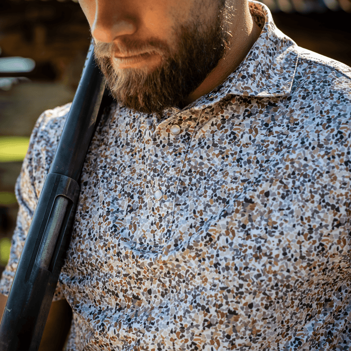 Man wearing the CamOAK Pearl Snap Polo by Iron Oak Apparel, featuring a unique camouflage pattern with earthy tones. He is holding a shotgun, capturing rugged style and Texan authenticity, perfect for outdoor adventures.