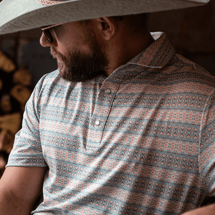 Man wearing The Desperado Pearl Snap Polo by Iron Oak Apparel, featuring a colorful horizontal Western pattern with shades of blue, orange, and white. He is seated, wearing a wide-brimmed hat and sunglasses, embodying a rugged yet stylish look.