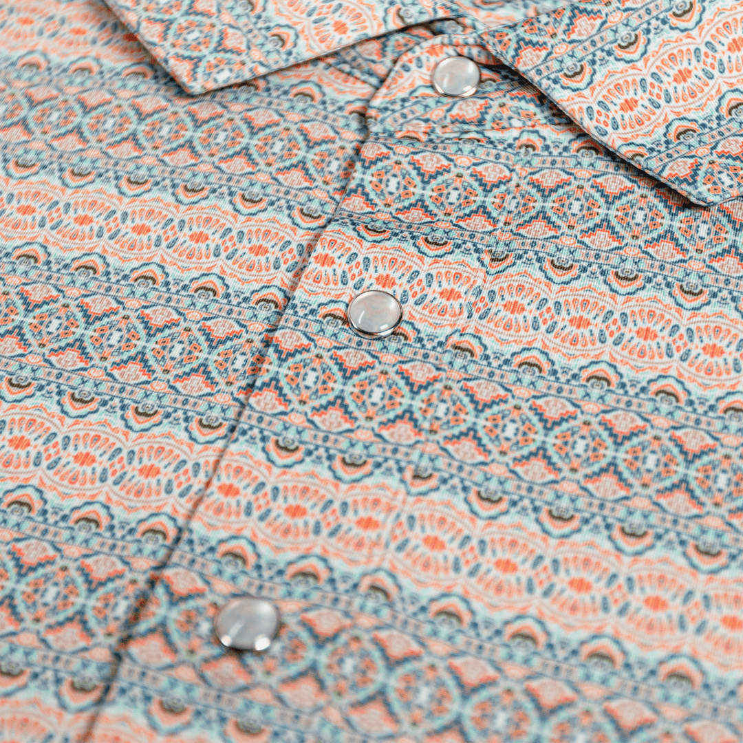 Close-up of The Desperado Pearl Snap Polo by Iron Oak Apparel, showcasing the intricate horizontal Western pattern with shades of blue, orange, and white and signature pearl snap buttons. This shirt adds a bold statement to any outfit.