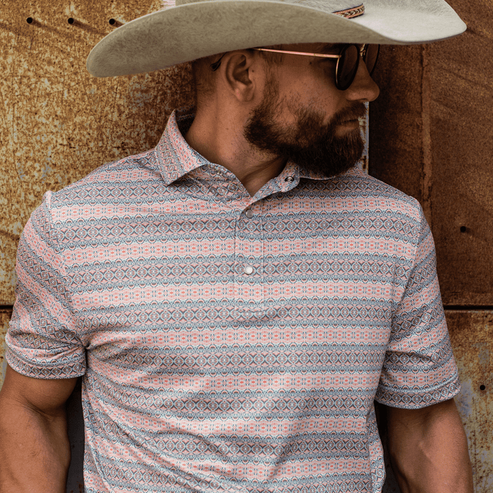 Man leaning against a rusted metal wall while wearing The Desperado Pearl Snap Polo by Iron Oak Apparel, featuring a colorful horizontal Western pattern with shades of blue, orange, and white. He is wearing a wide-brimmed hat and sunglasses, capturing a rugged yet stylish look.