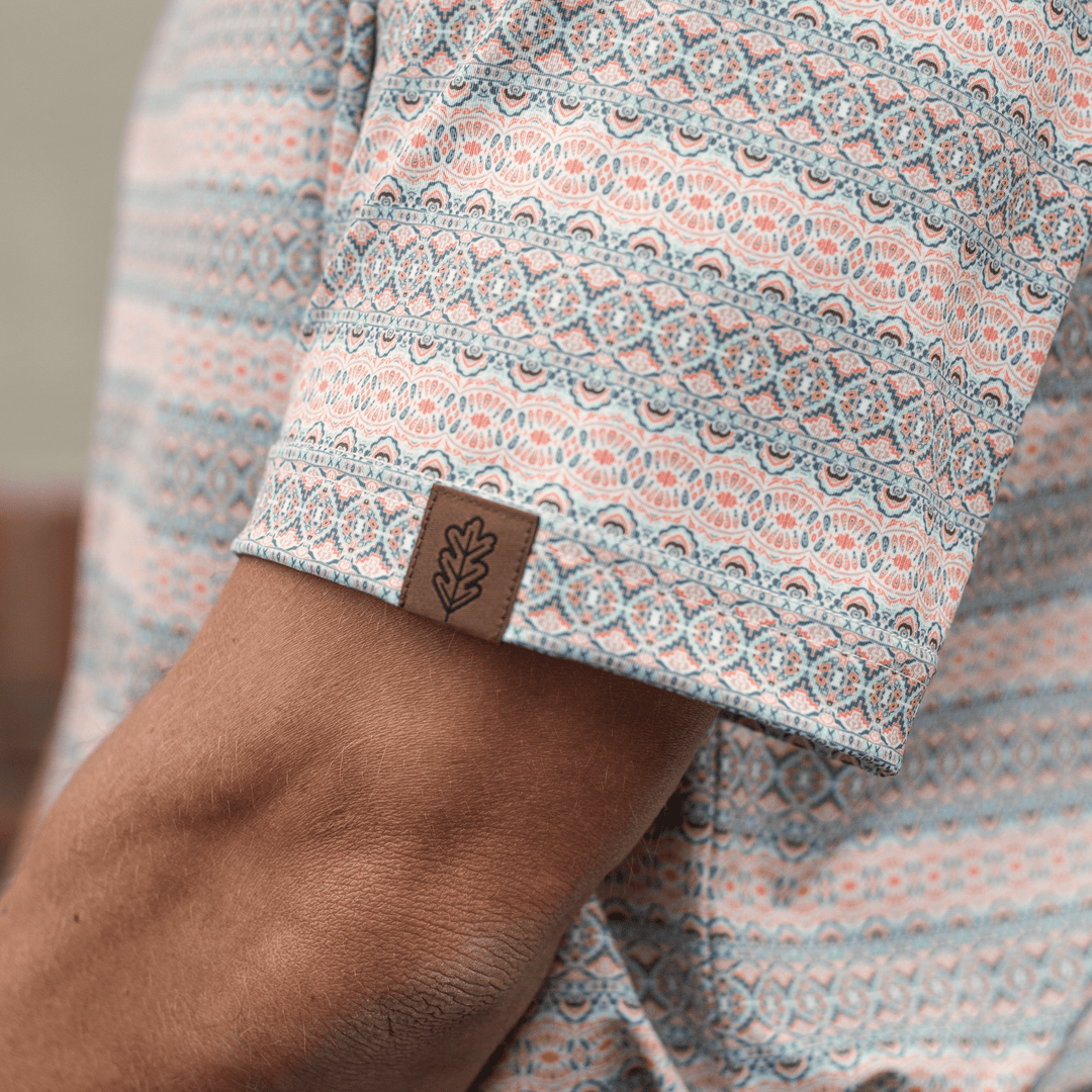 Close-up of the sleeve of The Desperado Pearl Snap Polo by Iron Oak Apparel, showcasing the intricate horizontal Western pattern with shades of blue, orange, and white and the brand's logo. This shirt is perfect for adding a bold statement to any outfit.