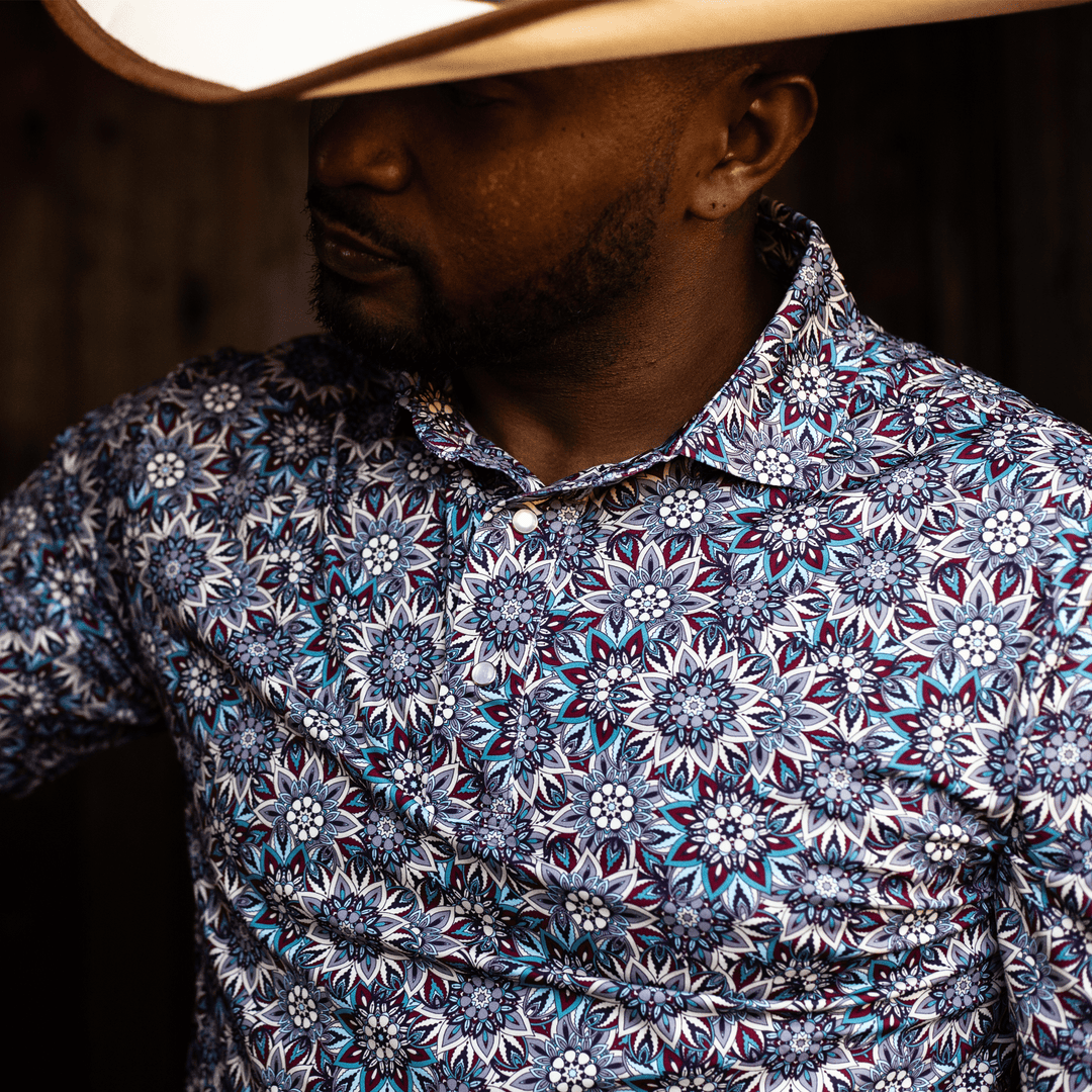 Man wearing The Hellbent Pearl Snap Polo by Iron Oak Apparel, showcasing the vibrant blue floral pattern. He stands indoors, exuding confidence and style.